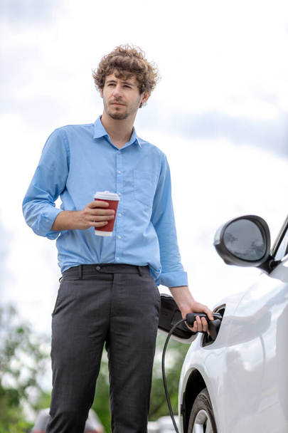 Progressive eco-friendly concept of parking EV car at public electric-powered charging station in city with blur background of businessman leaning on recharging-electric vehicle with coffee. - Foto, immagini