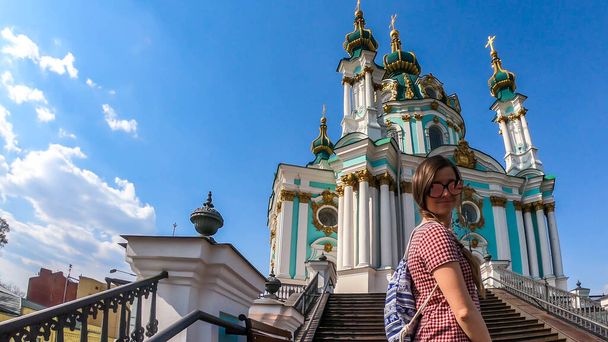 Молодая женщина в черном, стоящая на лестнице перед Андреевским храмом, Киев, Украина. Церковь относительно маленькая и окрашена в синий цвет. Чистое небо. Одинокая путешествующая девушка. - Фото, изображение