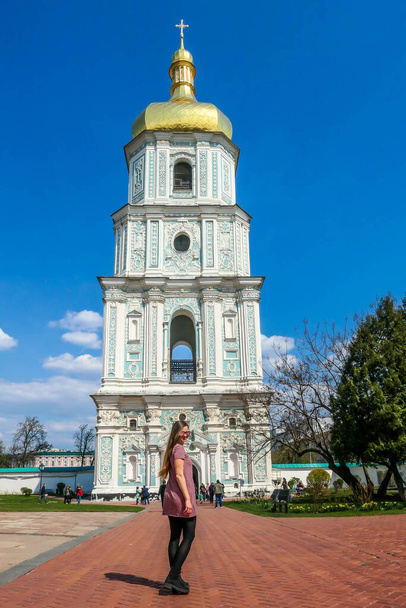 A young woman standing in front of St Sophia's Cathedral, Kiev, Ukraine. Golden rooftop of the tower. Play with the perspective, she seems as tall as the bell tower of the cathedral. Clear sky - Foto, afbeelding