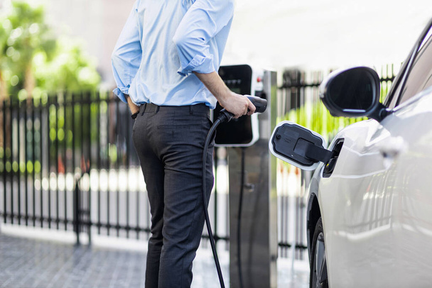 Closeup progressive man holding EV charger plug from public charging station for electric vehicle with background of residential building as concept eco-friendly sustainability energy car concept. - Foto, Imagen