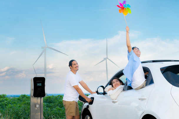 Концепція прогресивної щасливої сім'ї, що тримає вітряну іграшку і відпочиває на вітроелектростанції з електромобілем. Електричний транспортний засіб, керований чистою відновлюваною енергією від генератора вітрових турбін до зарядного пристрою
. - Фото, зображення
