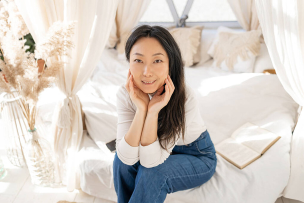 Jovem bela mulher asiática de meia idade sentada na cama em casa com luz natural. - Foto, Imagem