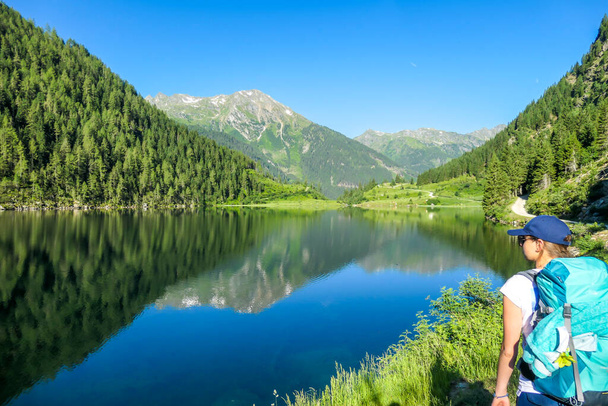 Молодая женщина с большим туристическим рюкзаком, стоящая рядом с чистым, синим озером, скрывающимся между высокими горными вершинами. Пышная зеленая трава и деревья, растущие на лугу. Весна в Альпах. - Фото, изображение