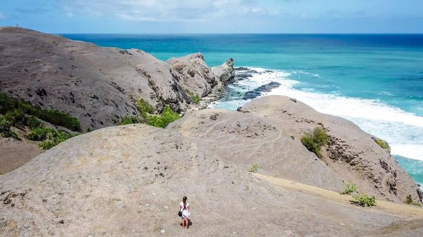 A woman in white dress standing on the cliffs, admiring the landscape in from of her. Overlooking famous Tanjung Aan beach on Lombok, Indonesia. Turquoise color of the water. The cliffs are barren. - Zdjęcie, obraz