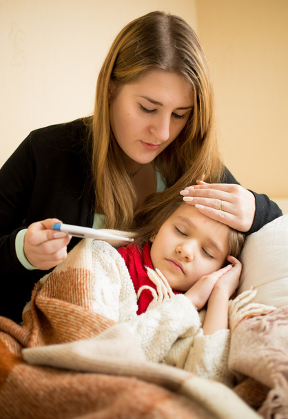 νέοι μητέρα αγκαλιάζει άρρωστος κόρη και τον έλεγχο θερμόμετρο  - Φωτογραφία, εικόνα