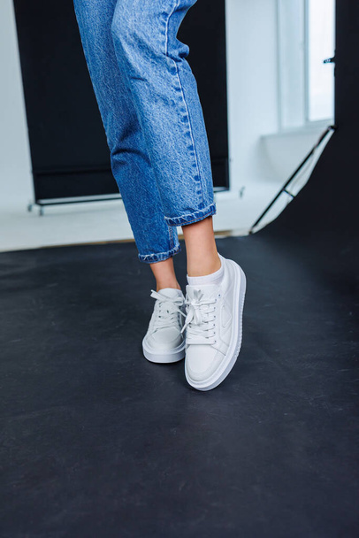 Close-up of female legs in jeans and casual white sneakers. Women's comfortable casual shoes. White leather women's sneakers - Fotó, kép