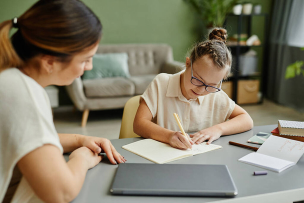 Portrait of young girl with down syndrome studying at home with female tutor or mother helping - Foto, Imagen