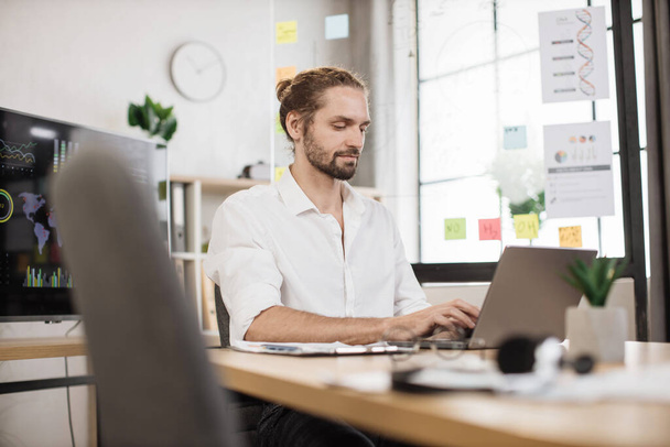 Portrait of attractive young business man sitting at office desk and working with laptop. Caucasian guy with beard having video conference. Business meeting online. - Fotó, kép
