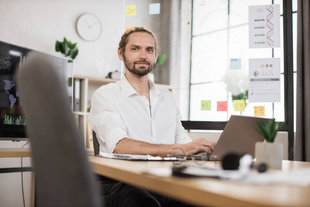 Business guy in stylish formal shirt posing at own modern office. Handsome young smiling bearded businessman sitting at desk with papers and modern laptop. - Fotografie, Obrázek