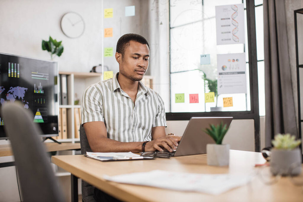 Busy african american man in striped shirt sitting at office and using wireless laptop. Big monitor with various charts and graphs on background. Business and finance concept. - Foto, Bild
