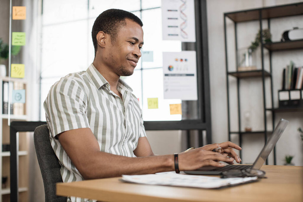 Portrait of attractive young businessman sitting at office desk and working with laptop. African american guy having online video conference. Business meeting online. - Foto, Imagem