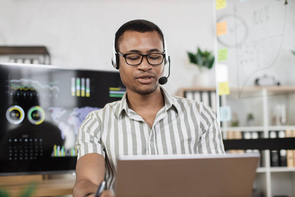 Confident african man in striped shirt using headset sitting at modern office and using wireless laptop. Huge monitor with various charts and graphs on background. Business finance technology concept - Fotografie, Obrázek