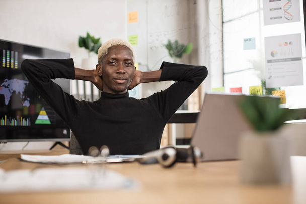 Впевнений африканський чоловік, який сидить розслабитися на робочому місці вдома, приймає сон або мрію про день. Щасливий розслаблений молодий чоловік відпочиває в кріслі відволікаючись від комп'ютерної роботи, знімає негативні емоції
. - Фото, зображення