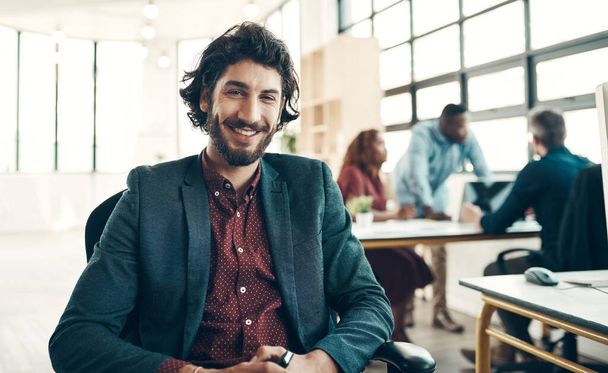 Portrait of a young businessman sitting in a modern office with his colleagues working in the background. - Photo, Image