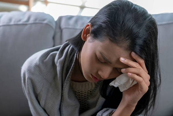Молодая азиатка, покрытая одеялом и страдающая головной болью от гриппа, сидит на диване, больная аллергическая девушка с симптомами аллергии кашляет дома - Фото, изображение