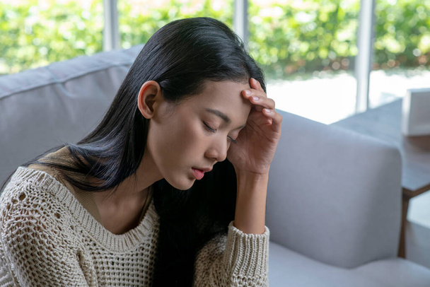 Päänsärky Nuori aasialainen nainen istuu sohvalla silmät kiinni, koskettaa päätään kärsiessään migreenistä.. - Valokuva, kuva
