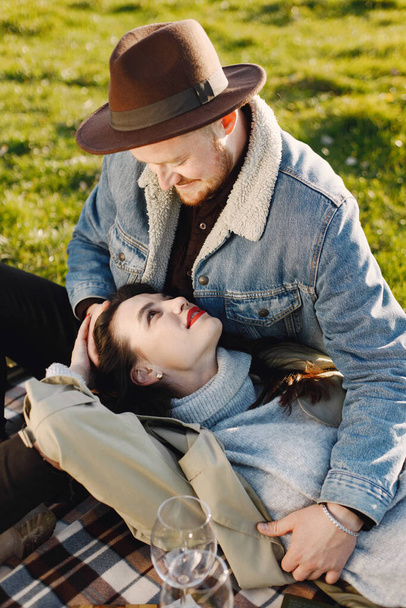 Man and woman in fashion clothes sitting on a nature on a picnic rug. Man wearing jacket and a black hat and woman coat. Woman lying on a mans lap. - Фото, зображення