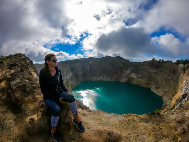 Frau sitzt am Vulkanrand und beobachtet die Kelimutu-Kraterseen in Moni, Flores, Indonesien. Die Frau ist entspannt und ruhig und genießt die Aussicht auf den See, der in vielen Türkistönen leuchtet - Foto, Bild