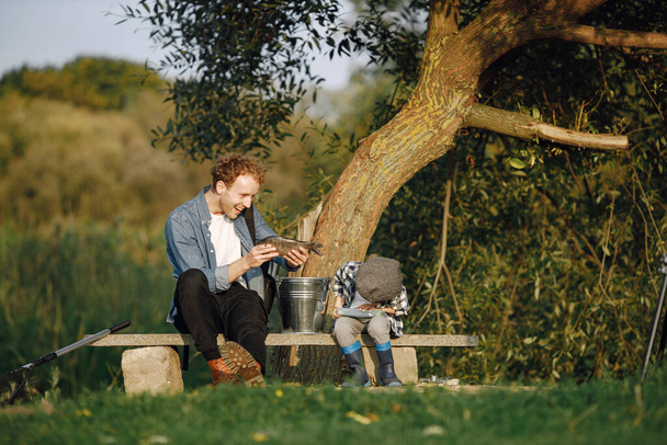 Young dad and his child boy spend time outdoors together. Curly toddler boy wearing a plaid shirt and a hat. Boy sitting near the tree and listening his father. - Photo, image