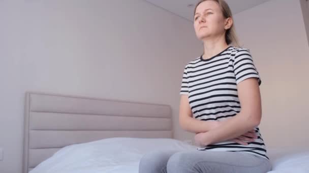 Mujer enferma que sufre sentada en la cama y sosteniendo el estómago. Una joven enferma. Dolor de estómago - Metraje, vídeo