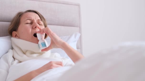 A young woman suffering from a sore throat lies on a beautiful bed and sprays her throat. Treatment of virus, cold and flu. - Imágenes, Vídeo