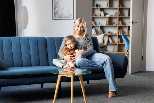 Young pregnant woman and her son sit on sofa at home, little boy spend time together, enjoy communication feeling kicks of unborn baby - Foto, Imagen