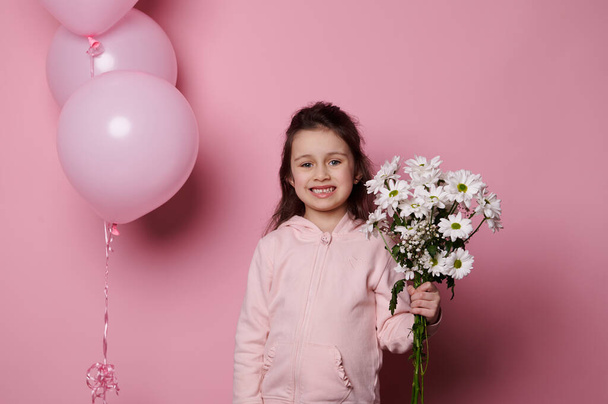 Smiling little girl, wearing pink pastel clothes, greeting with Womens or Mothers Day, standing near inflatable helium balloons, holding a bouquet of white spring flowers, isolated pink background - Fotografie, Obrázek