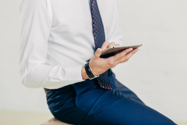 abgeschnittenes Bild eines erfolgreichen Geschäftsmannes in weißem Hemd, schwarzer Hose, Krawatte, hält ein modernes Tablet in der Hand, überprüft Benachrichtigung, ist mit drahtlosem Internet verbunden, isoliert über weißem Hintergrund - Foto, Bild