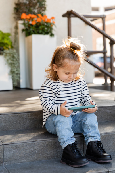 dziewczynka maluch w stroju casual za pomocą smartfona podczas siedzenia na schodach w pobliżu domu  - Zdjęcie, obraz