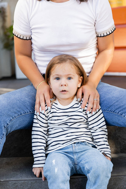 mãe em jeans sentado com a filha criança espantada no alpendre no quintal contra casa  - Foto, Imagem