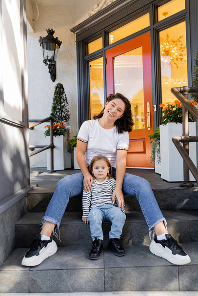 mère heureuse en jeans assis avec fille tout-petit sur le porche à l'arrière-cour contre la maison à Miami  - Photo, image