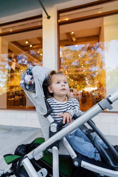 toddler girl in striped long sleeve shirt sitting in baby stroller near showcase of shop in Miami  - Foto, imagen