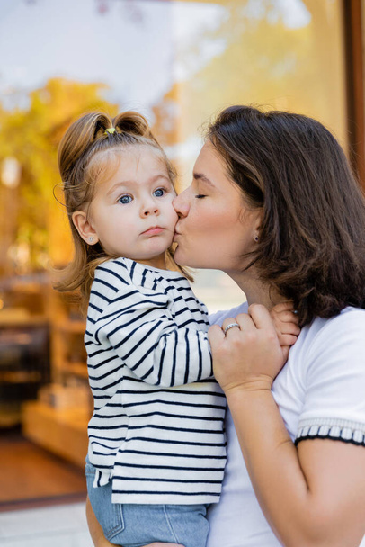 Брюнетка мать целует малышку в полосатой рубашке с длинным рукавом возле витрины магазина в Майами  - Фото, изображение