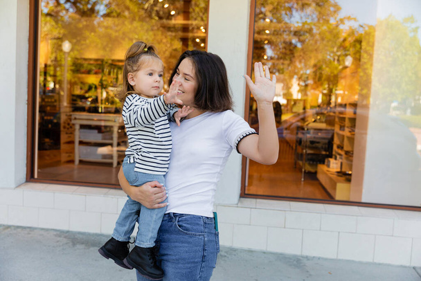 madre feliz sosteniendo en brazos bebé niña y haciendo gestos juntos cerca de escaparate de tienda en Miami  - Foto, imagen