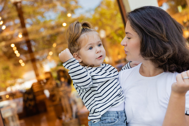 mère brune tenant dans les bras bébé fille en chemise rayée à manches longues près de vitrine de magasin à Miami  - Photo, image