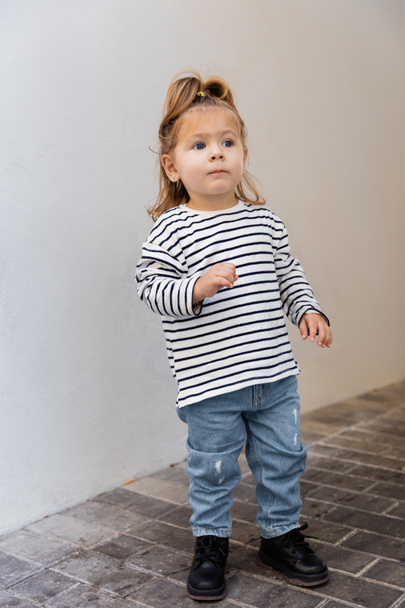 pełna długość malucha dziewczynka w paski koszuli z długim rękawem i dżinsy stojące w pobliżu białej ściany  - Zdjęcie, obraz
