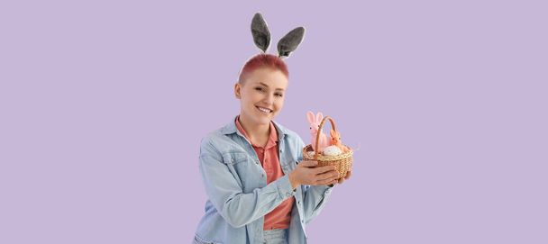 Young woman with basket of Easter eggs and bunnies on lilac background - Fotografie, Obrázek