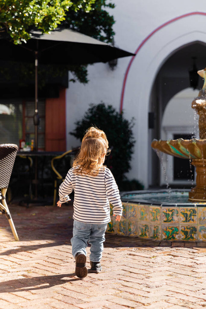 Rückansicht eines kleinen Mädchens in gestreiftem Langarmshirt und Jeans, das in der Nähe eines Brunnens in Miami spaziert  - Foto, Bild