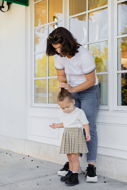 mother in jeans making hair of toddler daughter in skirt on porch of house  - Photo, Image