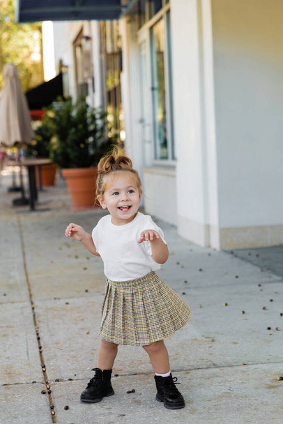 full length of positive toddler girl in skirt and white t-shirt standing on street in Miami  - Foto, afbeelding