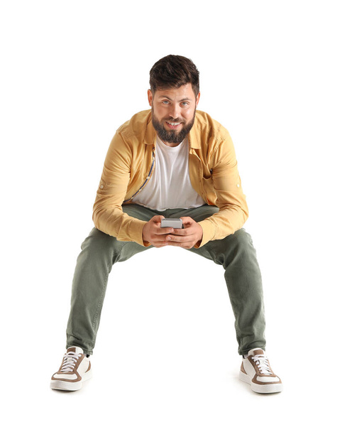 Young bearded man with mobile phone sitting in chair on white background - Photo, image