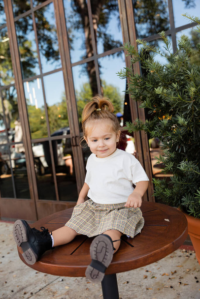 longitud completa de niña alegre en falda y camiseta blanca sentado en la mesa del bistro cerca de la cafetería al aire libre en Miami  - Foto, Imagen