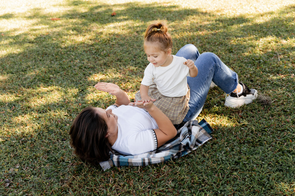 fröhliche Mutter liegt auf grünem Rasen und spielt Patty Cake mit fröhlichem Kleinkind Baby Girl in Miami Park  - Foto, Bild