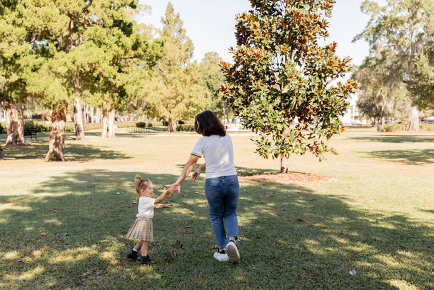 full length of brunette mother in jeans playing with happy toddler daughter in park of Miami  - 写真・画像