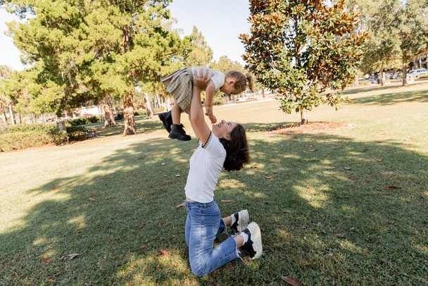 full length of cheerful mother in jeans lifting toddler daughter while playing in park of Miami  - 写真・画像