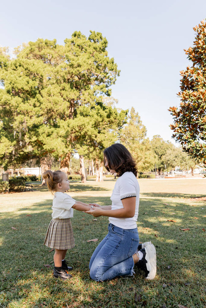 wesoła matka stojąca na kolanach i trzymająca się za ręce z córeczką podczas zabawy w parku Miami  - Zdjęcie, obraz