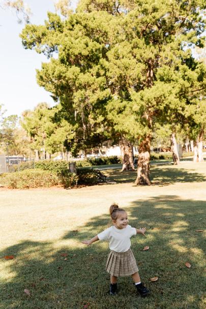 χαρούμενο κοριτσάκι στέκεται με απλωμένα χέρια ενώ παίζει στο πράσινο πάρκο  - Φωτογραφία, εικόνα
