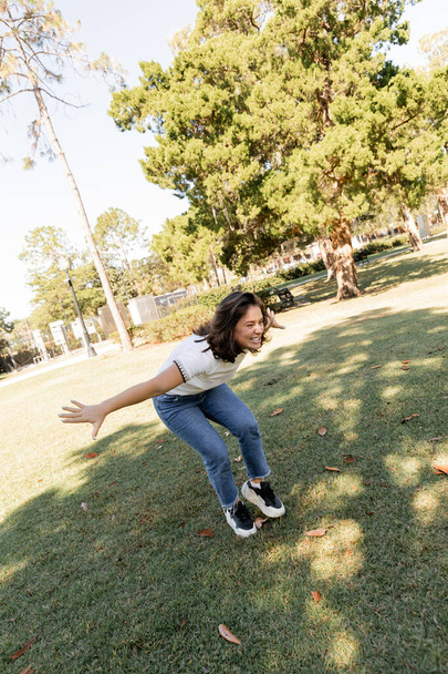mujer positiva en jeans de pie con las manos extendidas en el parque verde de Miami  - Foto, Imagen