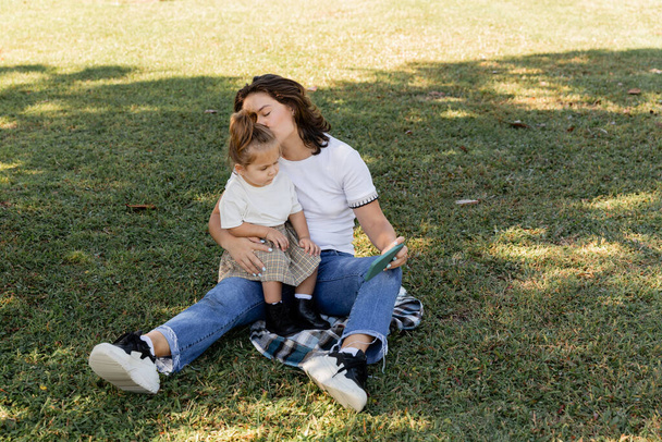 Mutter küsst Baby-Mädchen, während sie Smartphone in der Hand hält und auf Rasen sitzt  - Foto, Bild