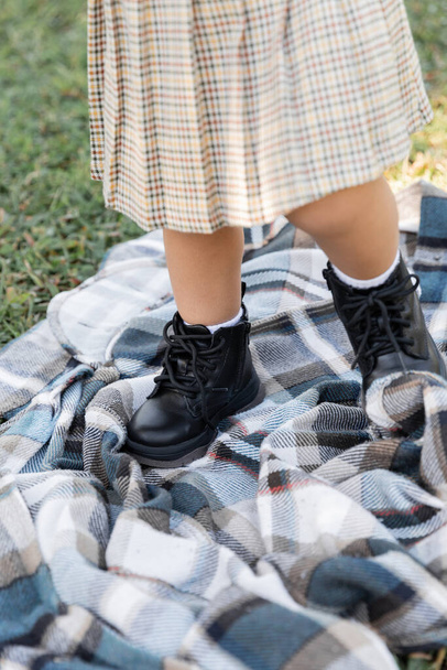 cropped view of baby girl in checkered skirt and boots standing on blanket near grass  - Фото, зображення